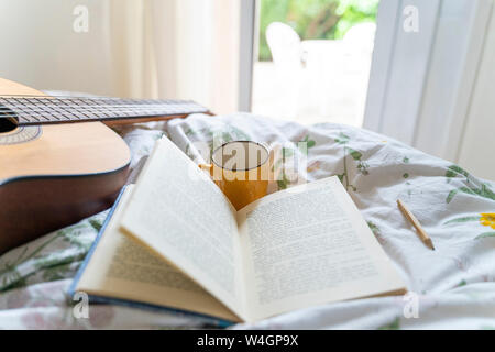 Guitar, coffee cup, pencil and open book on bed Stock Photo