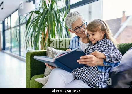 Grandmother sitting on couch with granddaughter, reading book together Stock Photo