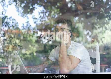 Young man with laptop on cell phone behind windowpane of a cafe Stock Photo