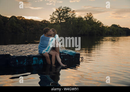 Young couple sitting on bathing platform, kissing, wrapped in towel Stock Photo