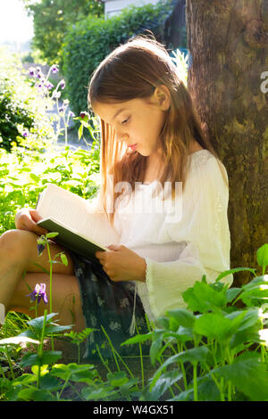 Little girl leaning against tree trunk reading a book