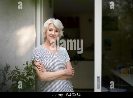 Portrait of smiling mature woman standing in front of open terrace door in the evening Stock Photo