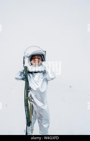 Smiling little girl wearing space suit putting on space hat in front of white background Stock Photo