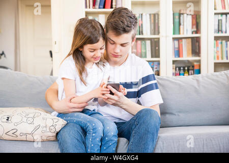 Teenage boy sitting with his little sister on the couch at home looking at cell phone Stock Photo