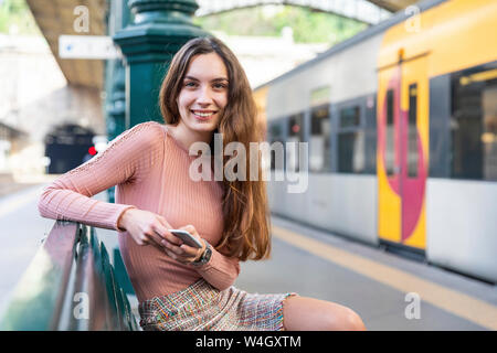 Portrait of smiling young woman with smartphone waiting on bench on platform, Porto, Portugal Stock Photo