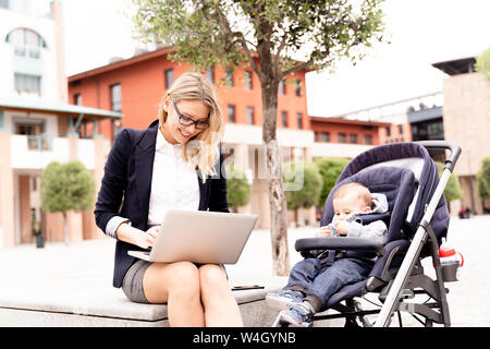 Young businesswoman with baby boy in stroller working on laptop outdoors Stock Photo