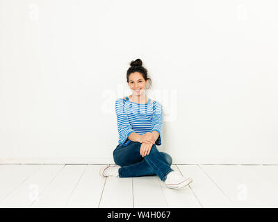 Beautiful young woman with black hair and blue white striped sweater sitting on the ground in front of white background Stock Photo