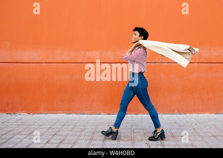 Businesswoman walking at a red wall in the background, putting her jacket on her shoulder Stock Photo