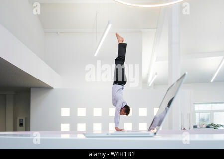Businessman doing a handstand at laptop in office Stock Photo
