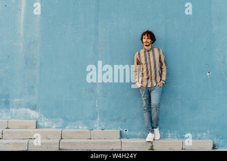 Bearded man wearing striped shirt leaning against light blue wall looking at distance Stock Photo