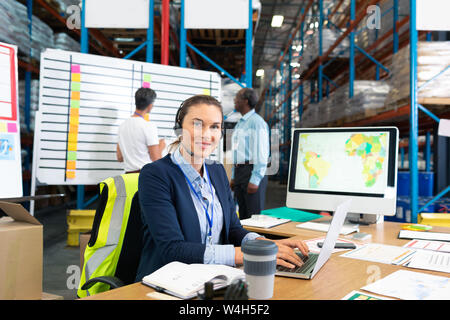 Female manager with headset using laptop at desk in warehouse Stock Photo