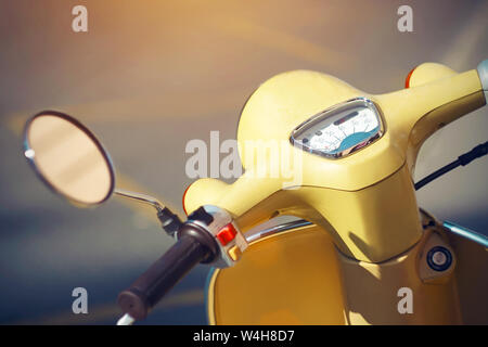 The steering wheel of a vintage yellow moped with a rear-view mirror and a speedometer, which stands on the road with yellow markings, illuminated by Stock Photo