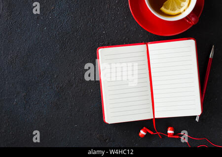 Blank red notebook with pen, headphones and cup of tea on dark table. Business still life, office or education concept. Top view of working desk. Stock Photo