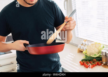 A young handsome brunette man is standing in the kitchen and holding a frying pan in his hands. Husband preparing breakfast for his wife. Stock Photo