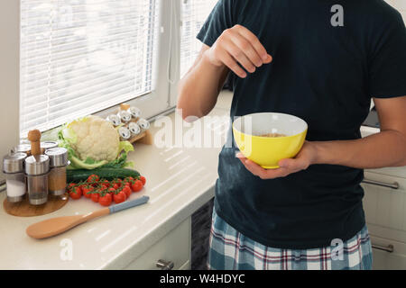 Young handsome man is having breakfast in the kitchen at home. The guy prepared himself oatmeal and a glass of milk for breakfast. Stock Photo
