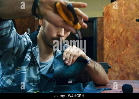 Young man sews leather goods. Craftsman creates leather object. Making handmade item. Guy working with needle pliers. Male hands with needle Stock Photo