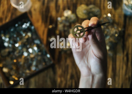 Watchmaker holds large gear. Cogwheel on blurred background of workplace. Pinion in female hand. Old cogwheel on wooden table background. Stock Photo