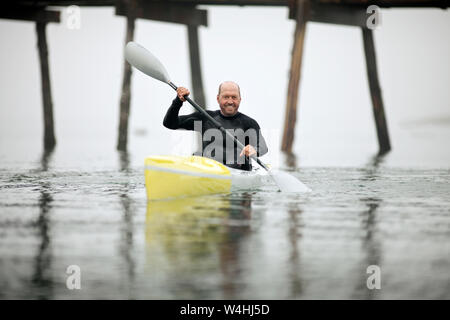 Male kayaker smiles for a portrait as he paddles in the waters of a foggy harbour. Stock Photo