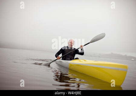 Male kayaker smiles for a portrait as he paddles in the waters of a foggy harbour. Stock Photo