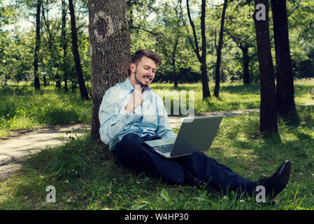 Young and happy businessman sitting on a ground with laptop in a park and making a successful gesture Selective focus Stock Photo