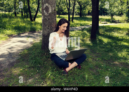 Young business woman sitting on a ground and working on her laptop and making gesture of success in a public park Selective focus Stock Photo