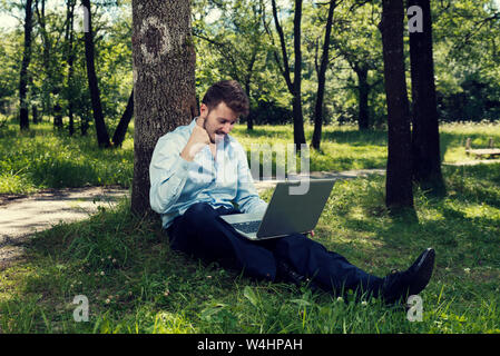 Young man working on his laptop in a public park sitting on the ground and making success gesture Selective focus Stock Photo