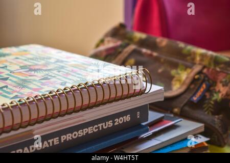 A pile of books, including a spiral notebook and a university prospectus, in a student's room, with a pencil case in the background. Stock Photo
