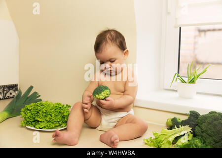 The baby in the diaper is sitting on the kitchen table. A child plays and has fun with fresh organic vegetables and fruits. Stock Photo