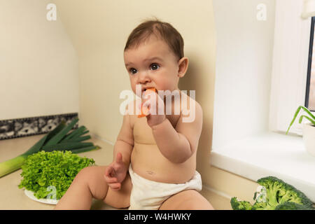 The baby in the diaper is sitting on the kitchen table. A child plays and has fun with fresh organic vegetables and fruits. Stock Photo