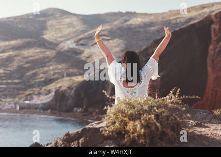 Back view of happy woman on the beach with hands up, sitting on rocks of famous red beach, with volcanic sand  and  rocky shoreline, Santorini, Akroti Stock Photo