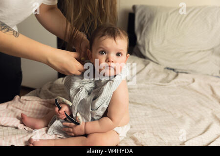 A young mother puts her baby in baby clothes. The baby obediently sits on the bed. Authentic lifestyle photos. Stock Photo