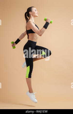A young attractive brunette woman with a ponytail is engaged in fitness, jumping with dumbbells, raising her knees high, on a peach background. Stock Photo