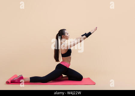 Young attractive girl with a ponytail, brunette, sitting in the pose of a pigeon with his hands on a pink yoga mat, on a pink background. Side view, Stock Photo