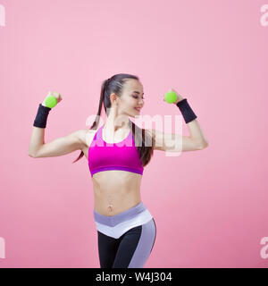 A young athletic brunette girl with a ponytail in a bright top shows off her biceps, holding a green dumbell in her hand on a pink background. Stock Photo