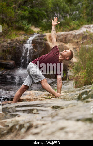 Man doing the triangle pose on rocks by a waterfall Stock Photo