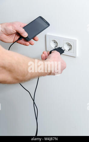 A man is inserting the plug into the wall socket to charge the smartphone. Stock Photo