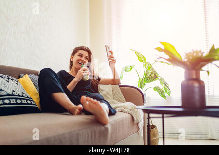 Happy young woman relaxing in living room and drinking smoothie using smartphone. Healthy diet Stock Photo