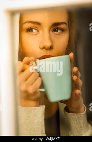 Young blond woman drinking coffee from cup and looking through the window. Stock Photo