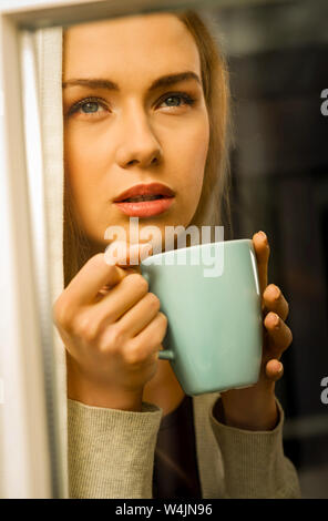 Thoughtful young blond woman with cup of coffee looking through the window. Stock Photo