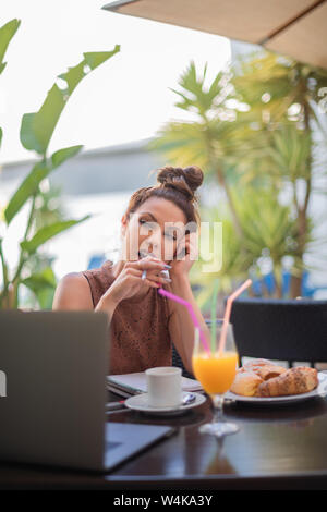 Beautiful young woman writer working on breakfast - Espresso coffee, orange juice, bread rolls and a laptop on the table - Portrait Stock Photo