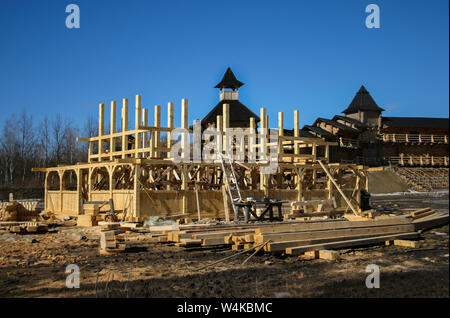 Construction of a traditional wooden house of uneven bars, logs and carved elements against the background of an ancient wooden fortress with towers. Stock Photo