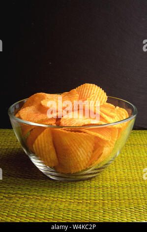 Fried chips in a transparent glass bowl, standing on a green wicker napkin on a black slate background Stock Photo