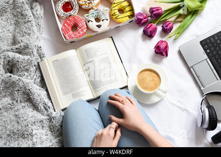 Girl reading a book and has brealfast in bed. Tray with strawberry pink donut with coffee and flowers. Doughnuts from food delivery. Top view. Stock Photo