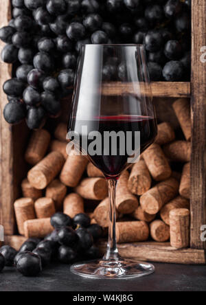 Elegant glass of red wine with dark grapes and corks inside vintage wooden box on black stone background. Stock Photo