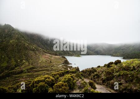 Aerial shot of a pond surrounded by green hills and forested mountain in a fog Stock Photo