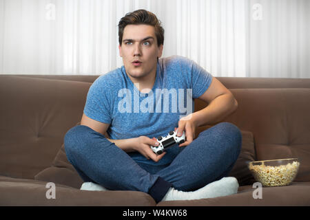 young handsome man playing video game sitting on comfy sofa holding joystick looks very concentrated on mission Stock Photo