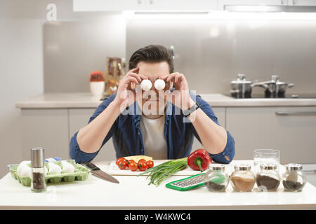young handsome man sitting in the kitchen preparing breakfast covers his eyes with fresh eggs having much fun Stock Photo