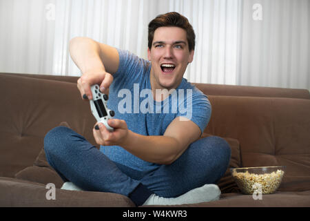 young handsome man playing video game sitting on comfy sofa holding joystick with both hands looking involved in mission Stock Photo