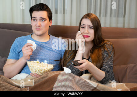 young couple watching movie on the sofa handsome man holding plate with popcorn and handkerchief attractive woman wiping tears away both looking Stock Photo