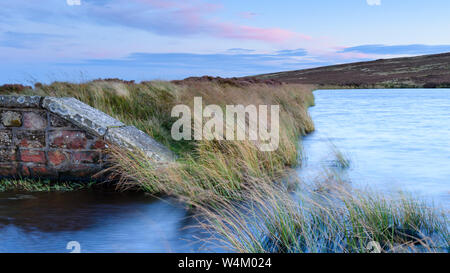 Lower Lanshaw Dam high on scenic moorland uplands (water, windblown grass, old brick wall, pink sunset sky) - Burley Moor, West Yorkshire, England, UK Stock Photo